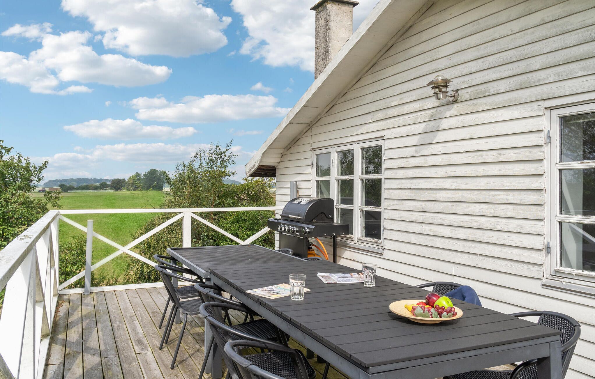 A wooden deck with a grill, outdoor table, and chairs, overlooking a green field.