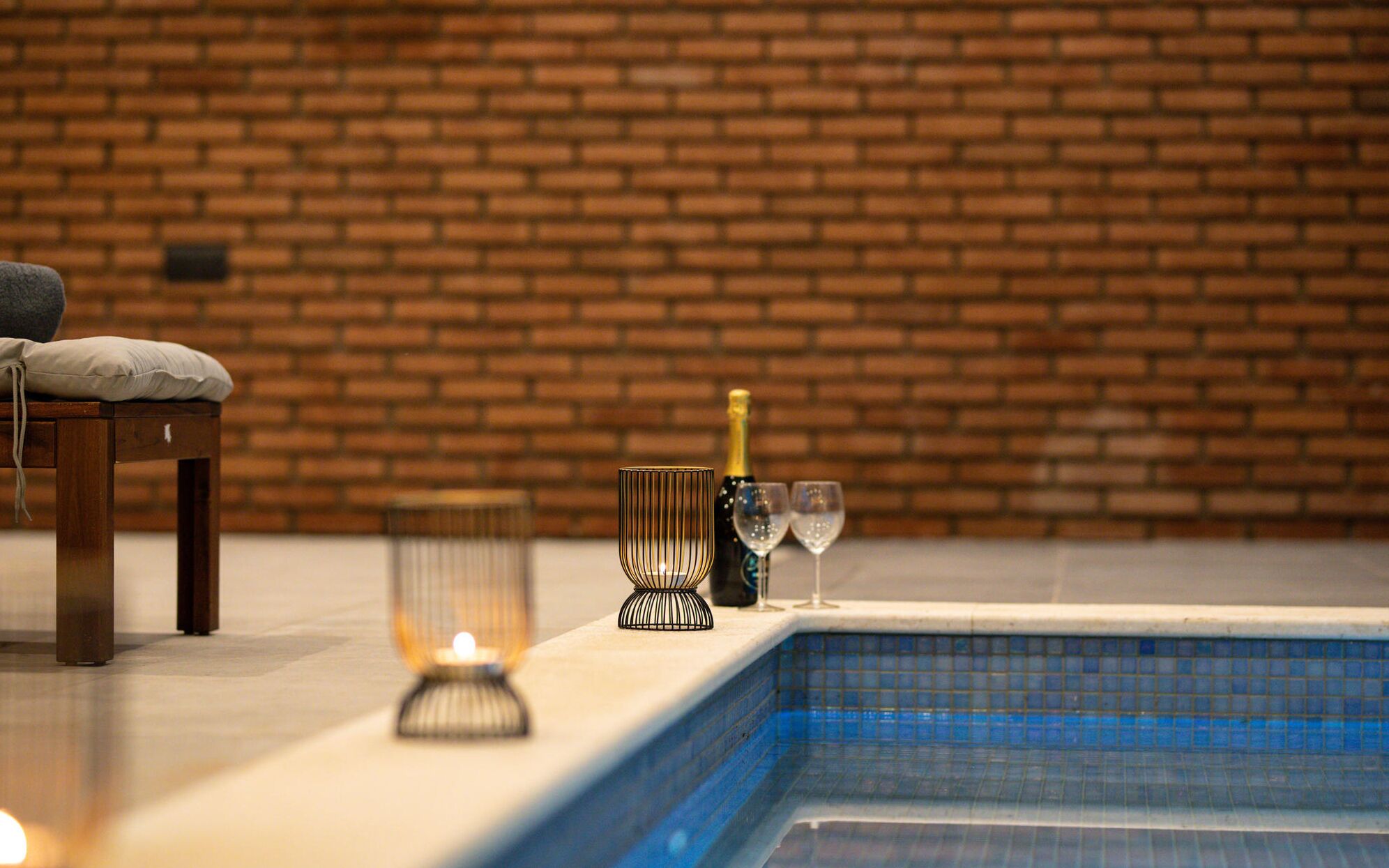 A poolside area with a brick wall, a cushioned chair, a candle, a bottle of champagne, and two glasses.
