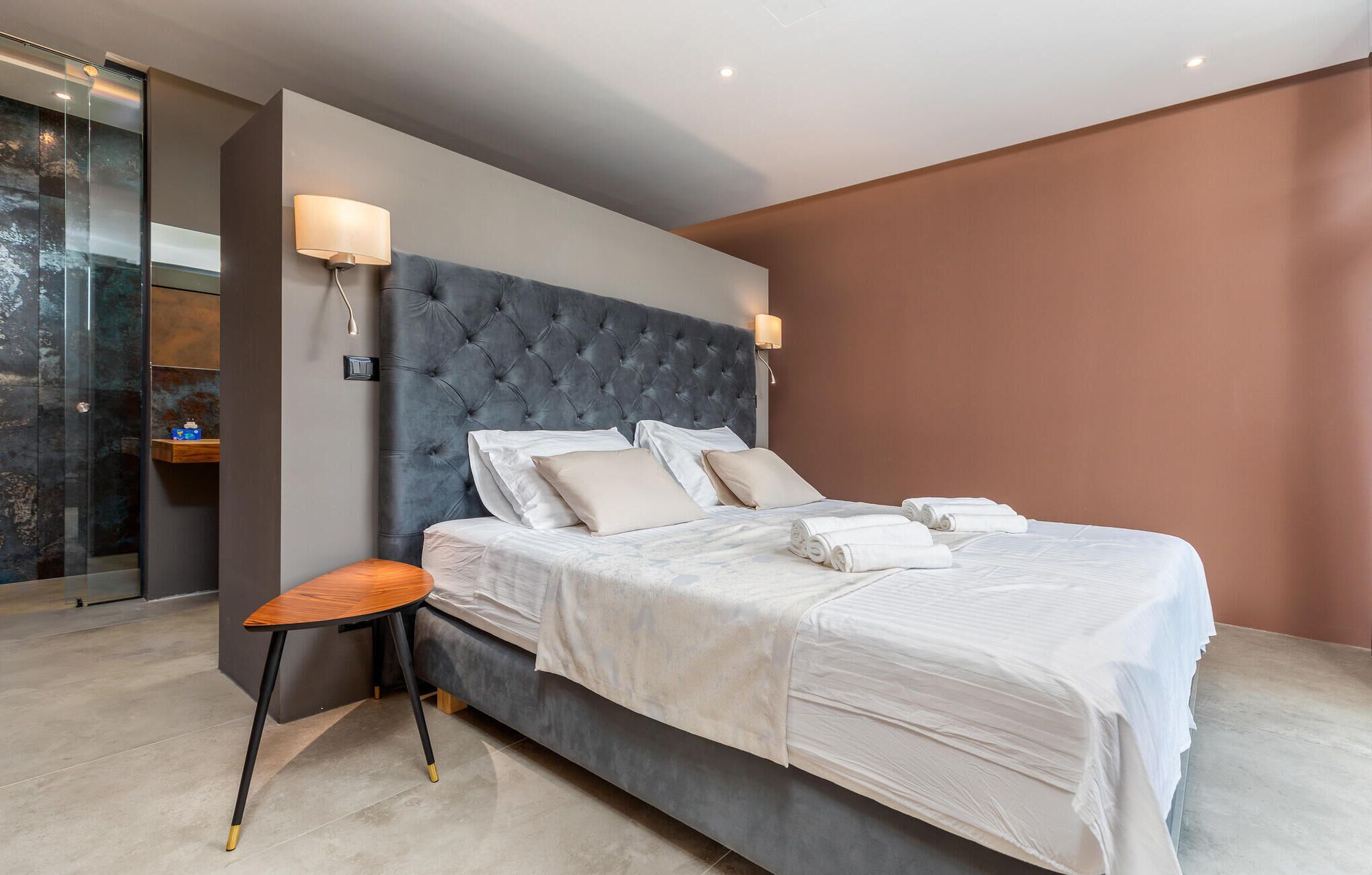 Une chambre à coucher moderne avec une tête de lit capitonnée, une table de chevet en bois et une cabine de douche en verre.