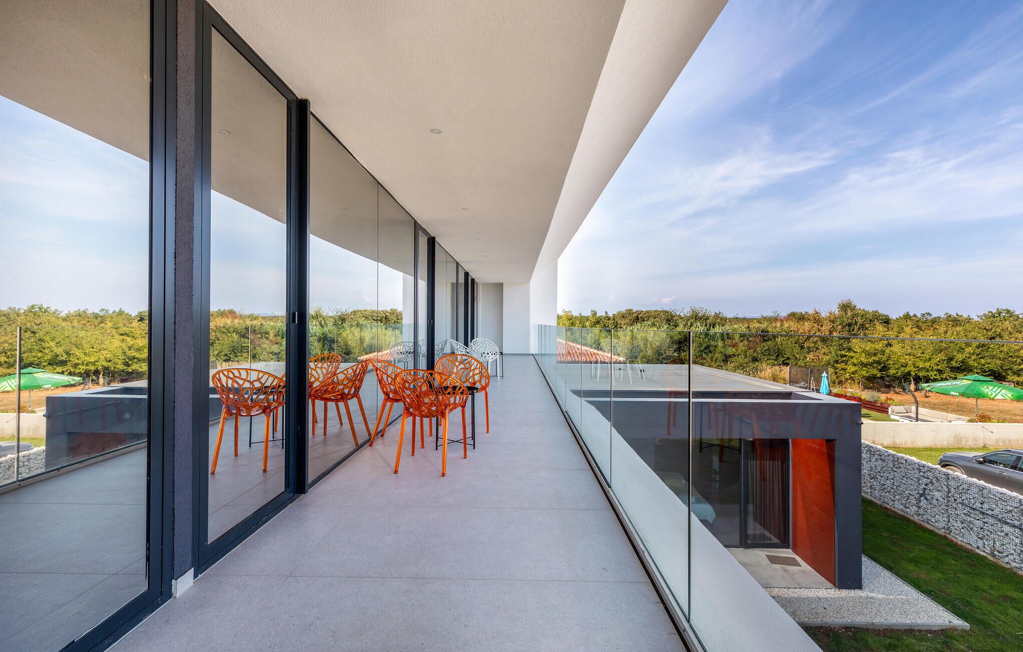 A modern balcony with glass railings, orange chairs, and a view of a green landscape.