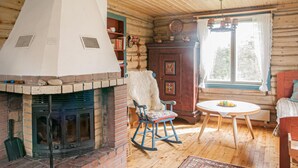 Interior rústico de cabaña con chimenea de piedra, paredes de madera, una mecedora, una mesa redonda y una ventana con cortinas.