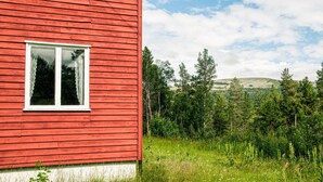 Un edificio de madera rojo con una ventana, rodeado de vegetación verde y una colina al fondo.