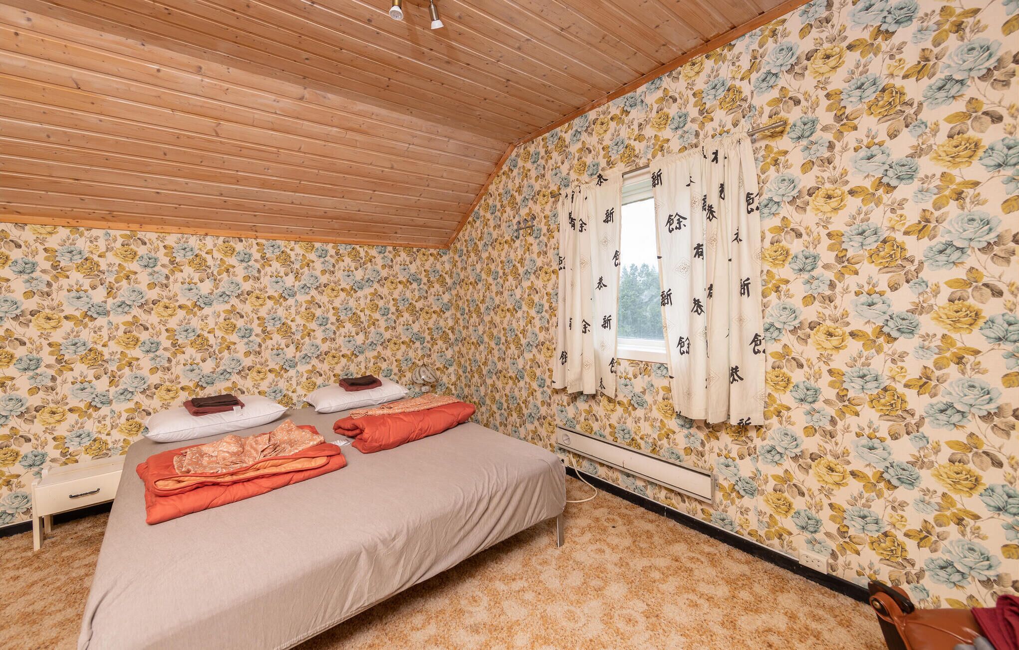 Une chambre à coucher avec un plafond en bois, un papier peint à motifs floraux, un lit avec des oreillers et des couvertures, une table de chevet et une fenêtre avec des rideaux.