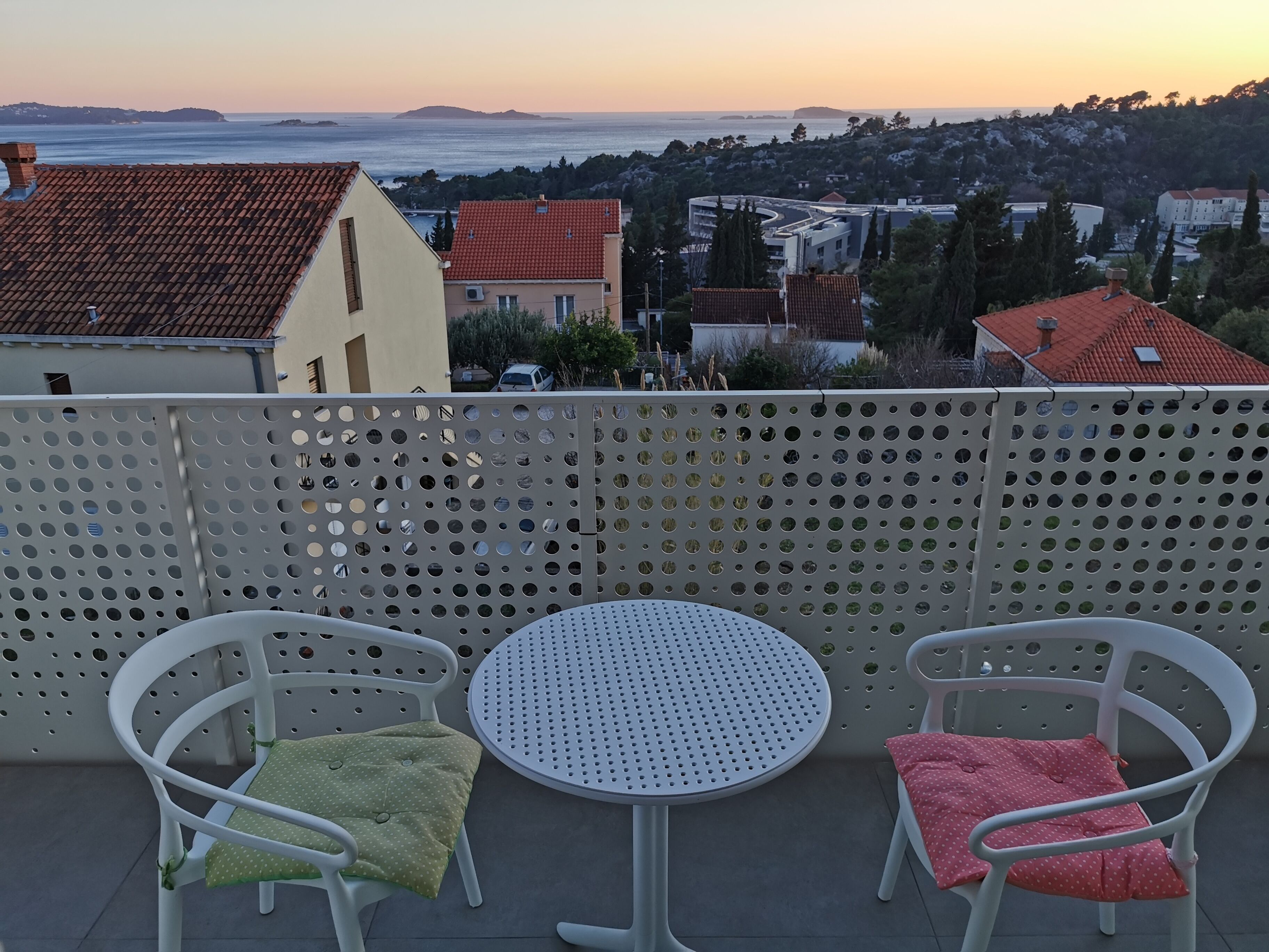 A balcony with two white chairs and a small round table, overlooking a coastal town with buildings and a view of the sea.