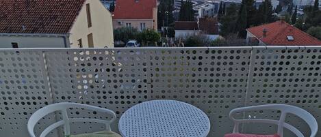 A balcony with two white chairs and a small round table, overlooking a coastal town with buildings and a view of the sea.