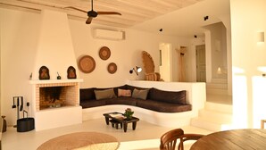 A living room with a fireplace, a sofa, a coffee table, and a ceiling fan. - Villa Santa Andrea with a fantastic 180 degree sea view (Mykonos)