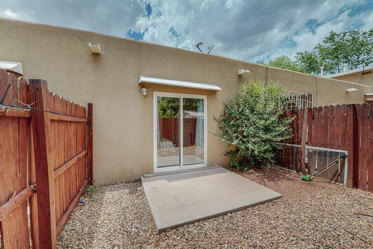 Un jardin à l’arrière d’une maison, avec une terrasse en béton, une clôture en bois et une porte-fenêtre coulissante donnant sur un espace de terrasse abritée.