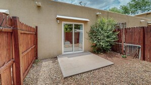 Un jardin à l’arrière d’une maison, avec une terrasse en béton, une clôture en bois et une porte-fenêtre coulissante donnant sur un espace de terrasse abritée.