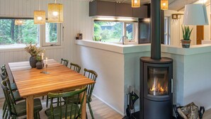Un comedor acogedor con una mesa y sillas de madera, una estufa de leña empotrada y un ventanal con vistas a un bosque.