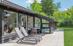 A covered outdoor seating area with lounge chairs and a table, overlooking a lush garden and a building with large windows.