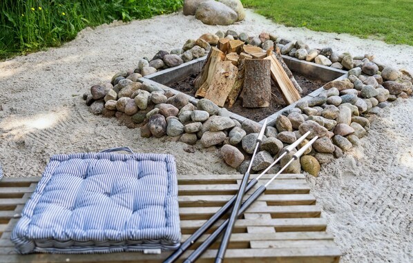A fire pit surrounded by rocks and logs, with a striped blanket and sticks on a wooden bench nearby.