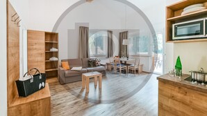 Un soggiorno moderno con divano, tavolino da caffè e zona pranzo. Si nota una libreria in legno e un angolo cottura con forno a microonde e una bottiglia verde.