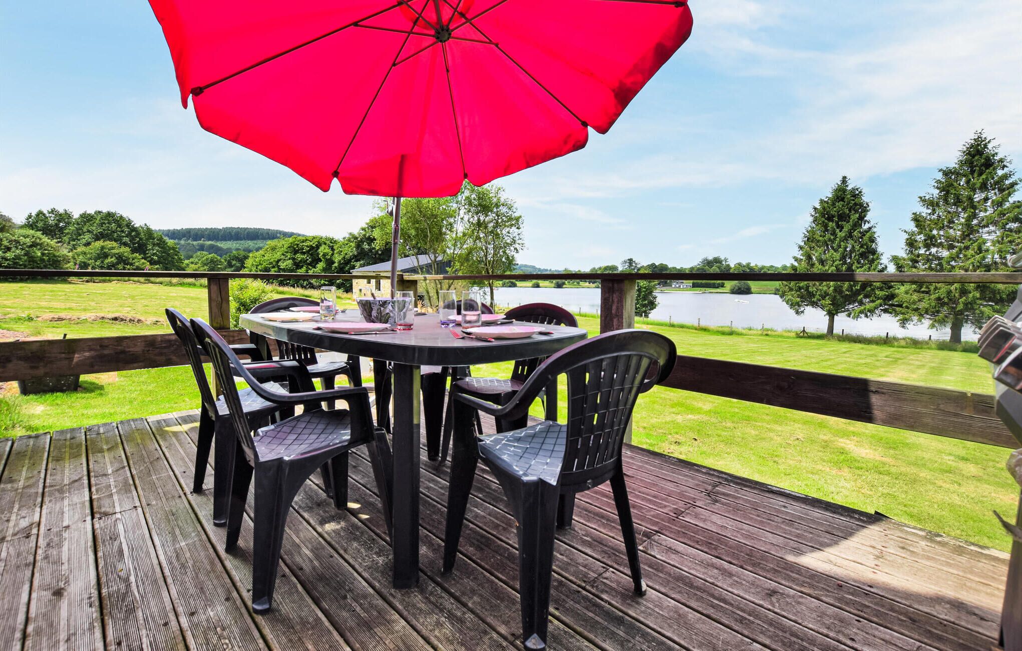 Eine Holzterrasse mit rundem Tisch und Stühlen, einem roten Sonnenschirm und Blick auf einen See und Bäume.