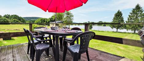 A wooden deck with a round table and chairs, a red umbrella, and a view of a lake and trees.