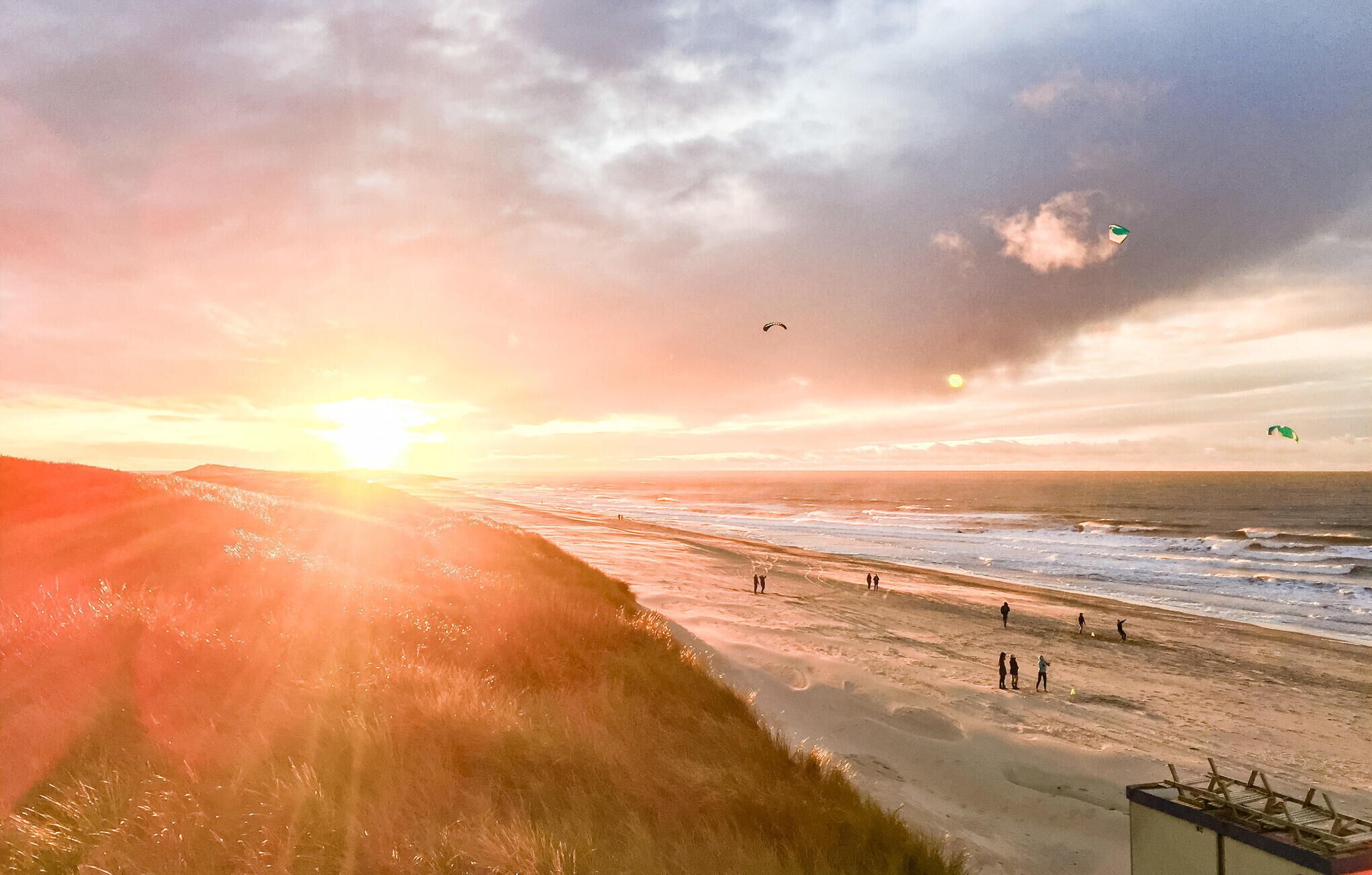 A beach scene with the sun setting over the ocean, people walking along the shore, and kites flying in the sky.