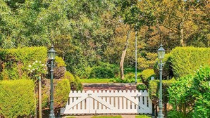 Un chemin de jardin mène à un portail blanc surmonté d’une clôture, le tout étant bordé de haies taillées et de lampadaires.