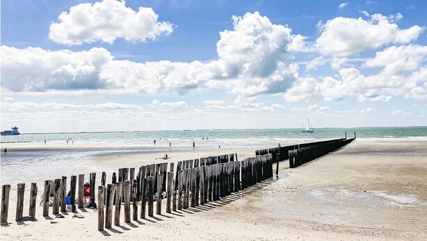 Une plage avec une jetée en bois, une mer calme et un voilier à l’horizon.