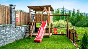 Una estructura de juego de madera con tobogán y zona de escalada, frente a una pared de piedra y rodeada por una valla de madera.
