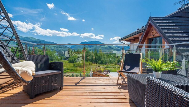 Una terraza de madera con mobiliario de mimbre, vistas a montañas y una barandilla de cristal.