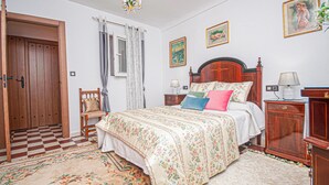 Un dormitorio con suelo a cuadros, una puerta de madera, una silla, una cama con ropa de cama floral, una mesita de noche, una lámpara y una pared con cuadros enmarcados.