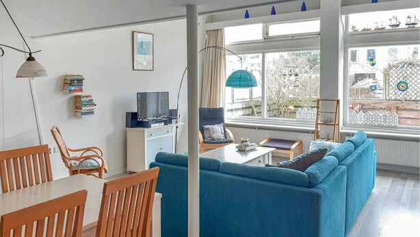 A modern living room with a blue sofa, wooden dining table, and chairs. There's a TV on a white cabinet, a bookshelf with books, and a window with a view of trees and buildings.
