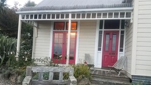 Una casa con un porche, dos puertas rojas, un banco de madera y un jardín con plantas y grava.