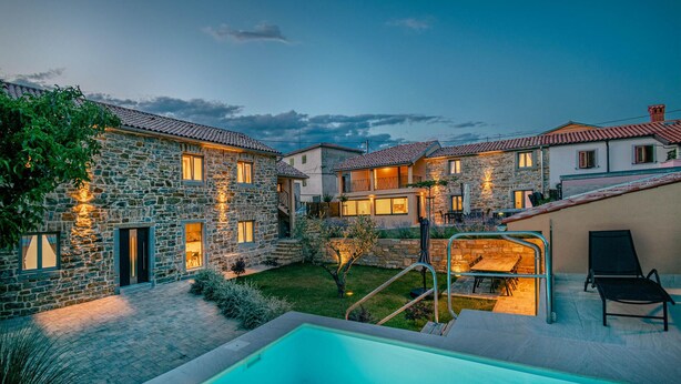 A stone villa with a swimming pool, outdoor seating, and a well-lit garden at dusk.