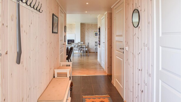 A hallway with wooden walls, a bench, a rug, and a dining area in the background.