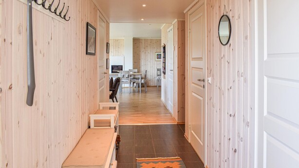 A hallway with wooden walls, a bench, a rug, and a dining area in the background.