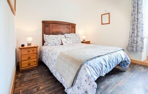 Une chambre à coucher avec une tête de lit en bois, un lit recouvert d’une literie à motifs floraux, deux tables de chevet avec des lampes, une commode en bois et une fenêtre avec des rideaux.