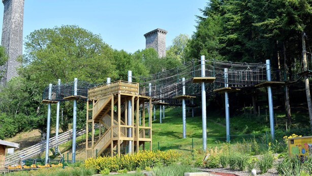 Eine Hängebrücke, Holzbauten und ein Steinturm in einem Park.