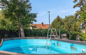 A swimming pool with a ladder, surrounded by trees and a building with a tiled roof in the background.