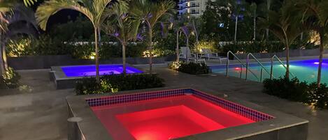Et udendørs poolområde om natten med et rødt oplyst boblebad, en blå oplyst rektangulær pool og liggestole.