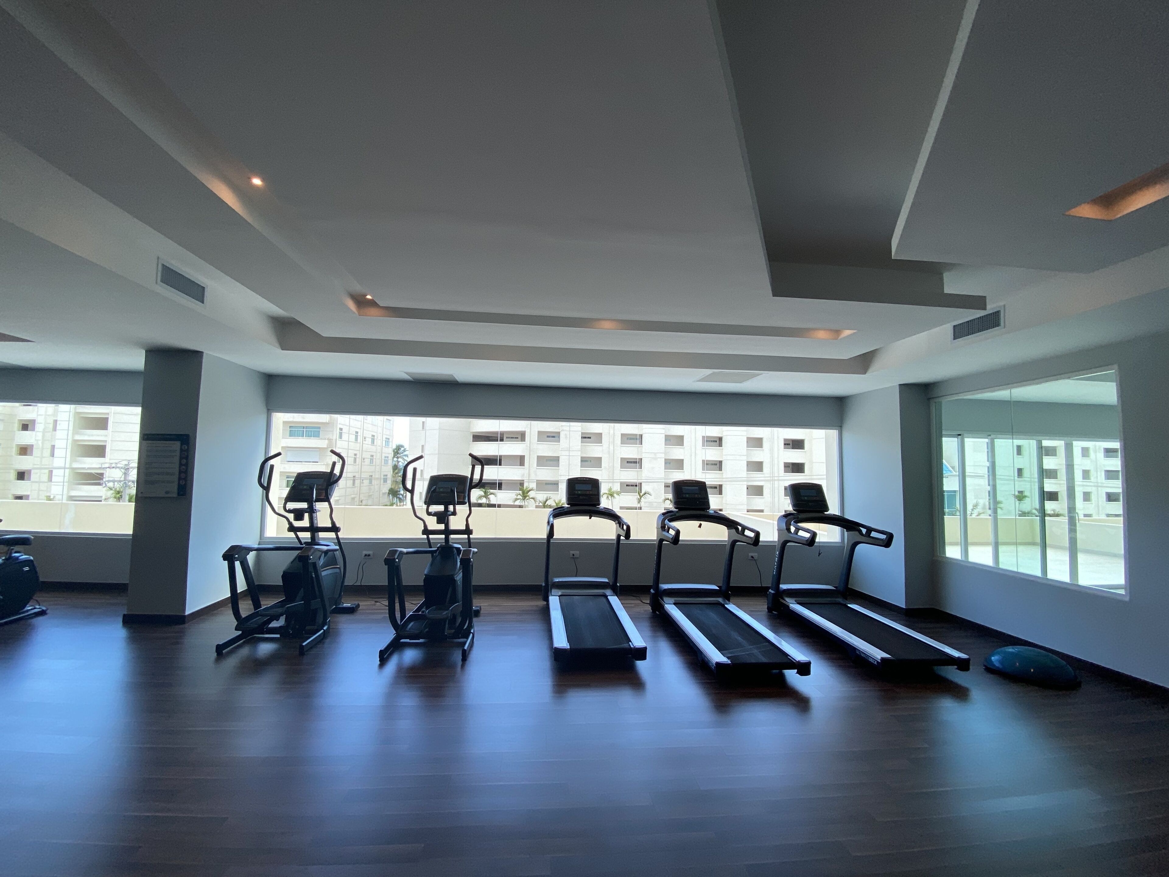 Ein Fitnessstudio mit Laufbändern, Crosstrainern und einem großen Fenster mit Blick auf ein Gebäude.