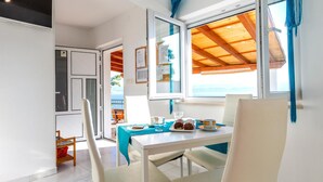 Un coin repas avec une table dressée pour deux personnes, une vue sur la mer par une fenêtre ouverte et un plafond en bois.