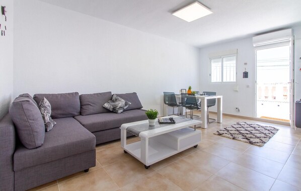 Ein modernes Wohnzimmer mit einem grauen Sofa, einem weißen Couchtisch mit Pflanze und einem Essbereich mit Glastisch und Stühlen.