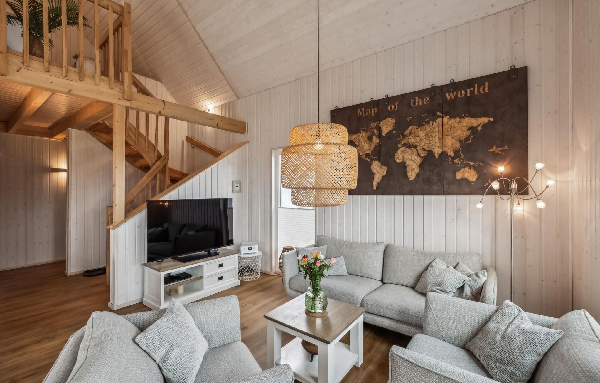 A modern living room with a wooden staircase, a large world map on the wall, a grey sofa, and a TV on a white cabinet.