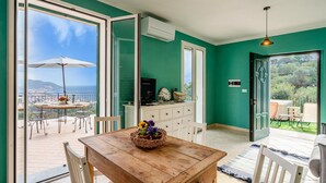 Ein Zimmer mit Holztisch, weißen Stühlen, einer Essecke mit Sonnenschirm, einem Fernseher und Blick auf das Meer.