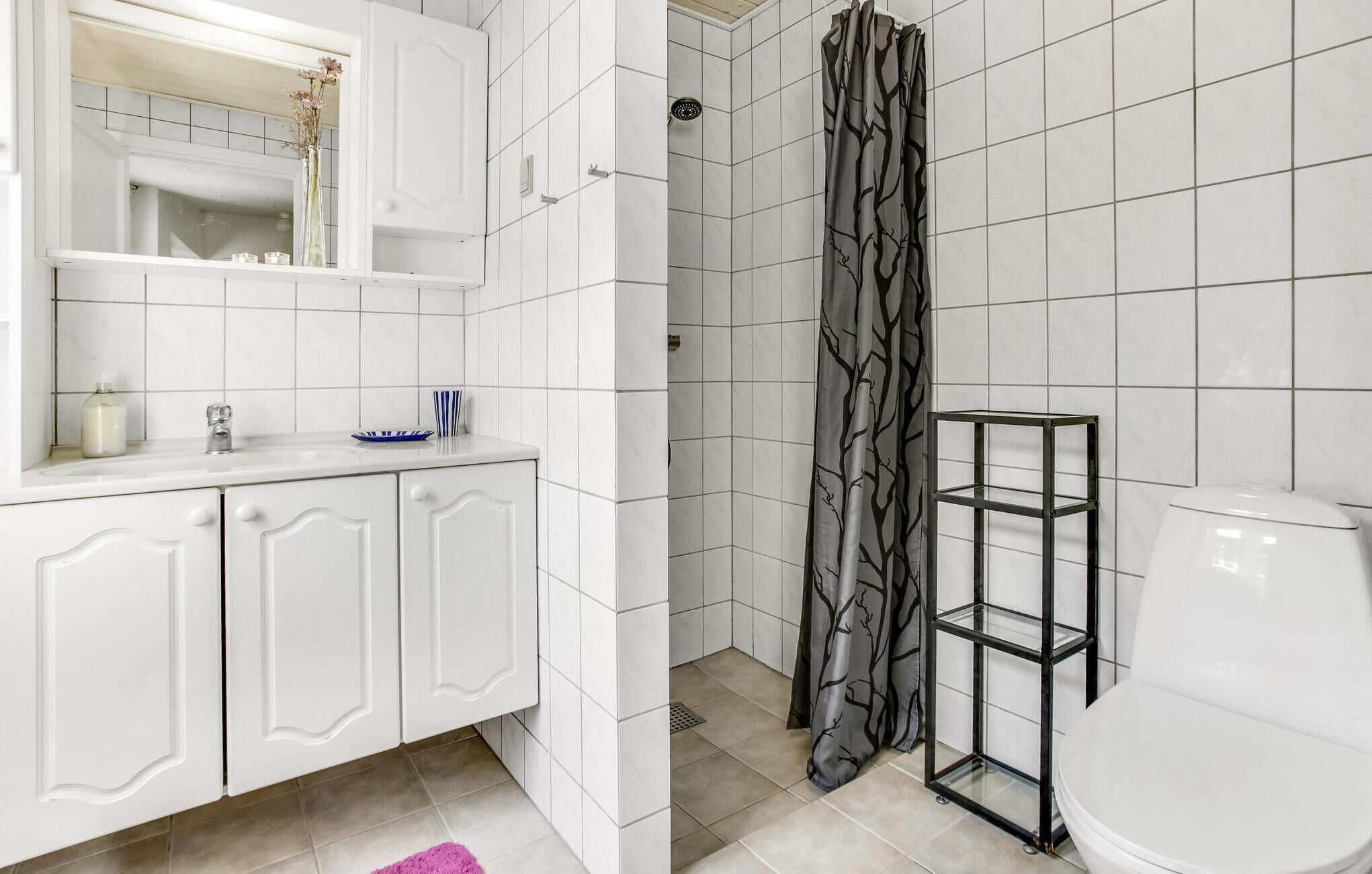 Ein Badezimmer mit weiß gefliesten Wänden, einer weißen Toilette, einer Dusche mit Vorhang mit Baummuster, einem weißen Waschbecken mit Spiegel darüber und einem rosa Teppichboden.
