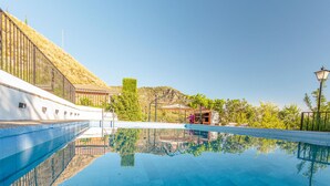 A clear swimming pool with a reflection of the surrounding landscape, a white building with a metal fence, and a hill in the background. - Beautiful home in Nigüelas (Nigüelas)