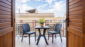 En åben balkon med to stole og et bord, en potteplante og udsigt til bygninger i baggrunden.