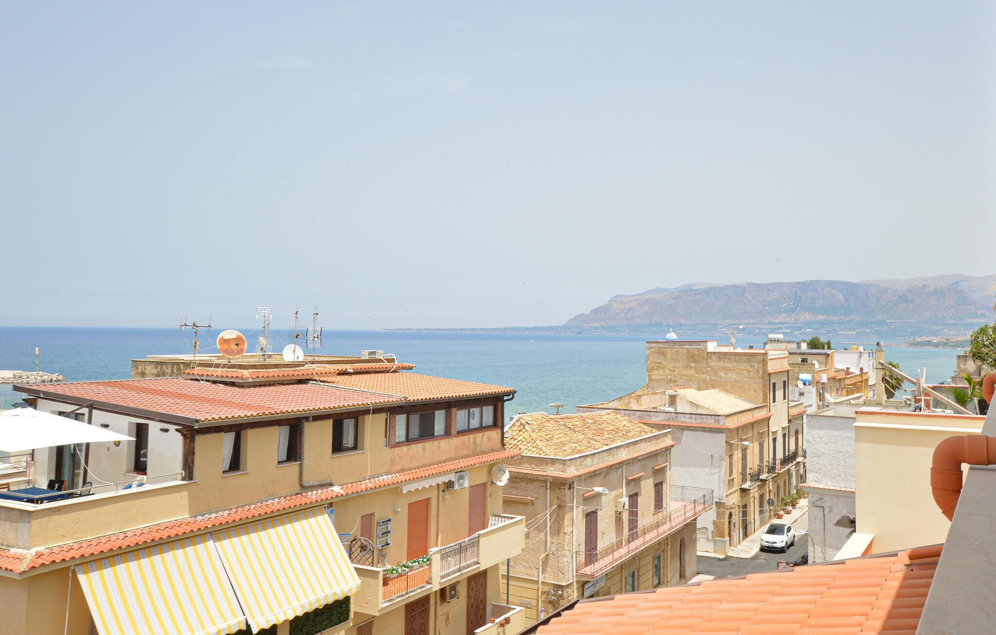 A coastal town with terracotta-roofed buildings, a clear view of the sea, and a distant mountain range.