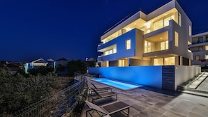 A modern two-story house with a swimming pool, outdoor seating, and a well-lit balcony.