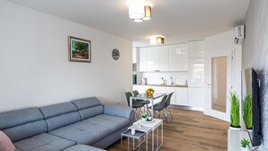 A modern living room with a grey sofa, a dining table with chairs, a kitchen area with white cabinets, and a TV on the right.