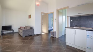 Ein modernes Wohnzimmer mit grauem Sofa, einem Flachbildfernseher und einem kleinen Tisch. Es gibt eine Küchenzeile mit weißen Schränken und einem Geschirrspüler. Der Raum hat einen Fliesenboden und eine Tür zu einem weiteren Zimmer.