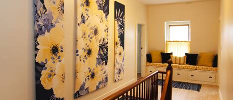 Un pasillo con tres grandes cuadros florales en la pared, una escalera con barandilla de madera y un rincón de descanso con sofá y cojines de diseño amarillo y negro.