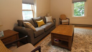 A living room with a sofa, a wooden coffee table, a chair, a small table with a lamp, and a window with a view of trees.