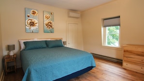 A bedroom with a large bed, two wall-mounted paintings, a bedside table with a lamp, a dresser, and a window with a view of trees.
