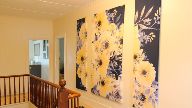 A hallway with a wooden staircase, a black and gold patterned rug, and three large floral paintings on the wall.