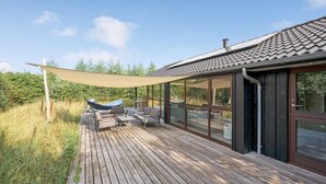 Una terraza de madera con mobiliario exterior, un toldo y una sala acristalada con techo de tejas.